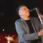 Fotografía de cuerpo entero del artista Jimmy Sax de pie tocando el saxofón con efecto de humo.