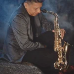 Jimmy Sax posando sentado con su saxofón dorado en una sesión de fotos nocturna con humo de fondo.